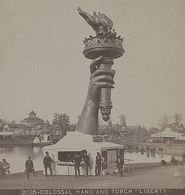 People stand on the torch held in the disembodied hand of the Statue of Liberty. Another group stands on the ground near an adjoining booth.