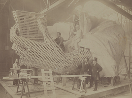 A group of men is assembled around the wood slats forming the unfinished arm of a plaster model of the Statue of Liberty.