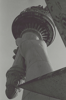 Closeup view of the Statue of Liberty’s hand holding the torch as seen through the spikes of the statue’s crown.
